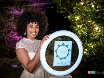 A smiling woman with curly hair stands next to a circular photobooth device lit up with a ring light. She is wearing a white, lace dress and has her hand on the photobooth. The background is decorated with lights and greenery, showcasing the elegance of l