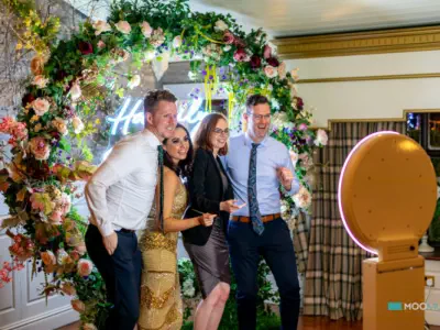Four people are posing in front of a floral backdrop with a neon sign that reads "Happily Ever After." They are smiling and looking at the camera of their digital photo booth hire in Wiltshire. The backdrop is beautifully adorned with pink, white, and gre