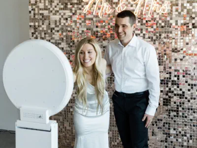 Three people stand around a digital photo booth hire in Gloucestershire, illuminated by a circular ring light. Two women, one in a blue dress and the other in a red dress, interact with the booth, while an older man in a suit watches. The background featu
