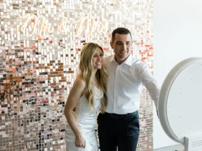 Two young women pose playfully for a photo booth in a lively bar environment. They lean in close together, with animated expressions on their faces. The bar behind them is bustling, with patrons sitting and standing near the counter.