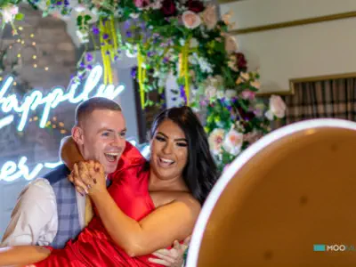 A man in a blue vest and white shirt happily carries a woman in a red dress. They smile joyfully at a photo booth, with a neon "Happily Ever" sign and a vibrant floral arch in the background.