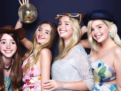 A digital illustration of four smiling young women with shiny hair. Each woman holds a brightly colored plush toy. They are grouped closely together, appearing cheerful and happy against a dark background. The overall mood is joyful and fun.