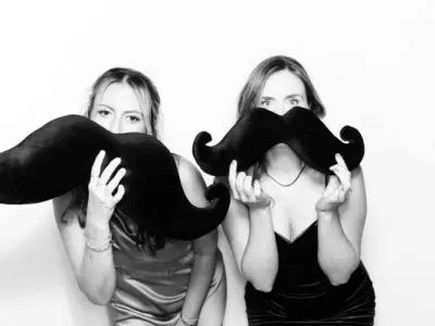 A smiling woman with curly hair stands next to a circular photobooth device lit up with a ring light. She is wearing a white, lace dress and has her hand on the photobooth. The background is decorated with lights and greenery, showcasing the elegance of l