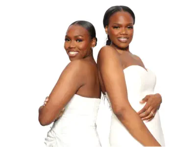 Three people stand around a digital photo booth hire in Gloucestershire, illuminated by a circular ring light. Two women, one in a blue dress and the other in a red dress, interact with the booth, while an older man in a suit watches. The background featu