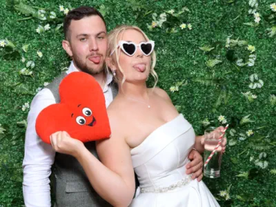 Two young women pose playfully for a photo booth in a lively bar environment. They lean in close together, with animated expressions on their faces. The bar behind them is bustling, with patrons sitting and standing near the counter.